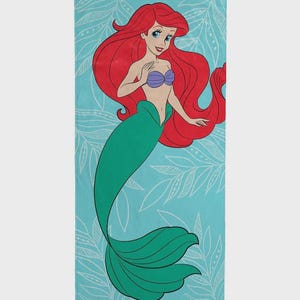 Op de afbeelding: Een rechthoekige strandlaken met een cartoonillustratie van een zeemeermin met lang rood haar, een paarse schelp top en een groene staart. De zeemeermin staat tegen een lichtblauwe achtergrond met witte bladerpatronen.