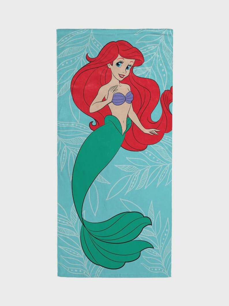 Op de afbeelding: Een rechthoekige strandlaken met een cartoonillustratie van een zeemeermin met lang rood haar, een paarse schelp top en een groene staart. De zeemeermin staat tegen een lichtblauwe achtergrond met witte bladerpatronen.