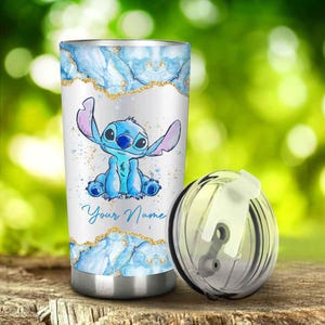May include: A stainless steel tumbler featuring a blue cartoon character with large ears. The tumbler has a white background with blue and gold accents. The words "Your Name" are written in a script font. A clear lid sits next to the tumbler.