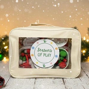 Könnte beinhalten: Cremefarbene Stofftasche mit transparentem Fenster, das farbenfrohes Bastelzubehör zeigt. Die Tasche hat einen Griff und ein rundes Etikett mit der Aufschrift "Seasons of Play". Inhalt: rote und grüne Pompons und andere Bastelartikel.