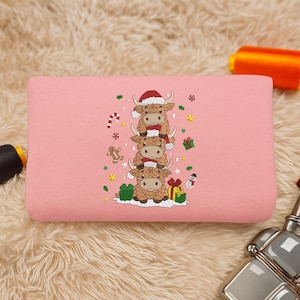 May include: A pink rectangular cushion with an embroidered design of three stacked Highland cows wearing Santa hats and scarves. The cows are surrounded by Christmas-themed elements like snowflakes, gifts, and a gingerbread man.