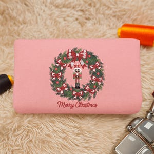May include: A rectangular, light pink pillow with a Christmas wreath design. The wreath features red bows, green leaves, and a nutcracker in the center. The words "Merry Christmas" are written below the wreath.