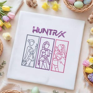 Puede incluir: Camiseta blanca con la palabra "HUNTRX" en morado, encima de tres ilustraciones enmarcadas de figuras estilizadas. Las ilustraciones están delineadas en gris, morado y rosa. Decoraciones de Pascua rodean la camiseta.