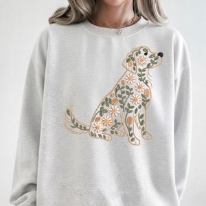 Könnte beinhalten: Hellgraues Sweatshirt mit floralem Hundedesign. Der Hund sitzt und ist mit weißen und gelben Blumen, grünen Blättern und beigen Akzenten verziert. Das Sweatshirt hat einen Rundhalsausschnitt und lange Ärmel.