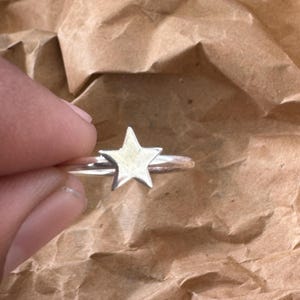 May include: A silver ring with a five-pointed star design. The star is the focal point on a slender band. The ring is set against a brown, textured backdrop.