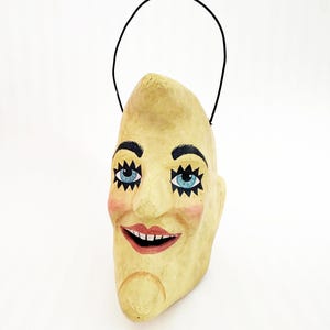 May include: A quirky, hand-painted paper mache mask shaped like a crescent moon with a smiling face. It has blue eyes, black eyebrows, rosy cheeks, and a red smiling mouth. A black wire handle is attached.