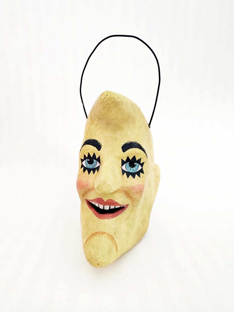 May include: A quirky, hand-painted paper mache mask shaped like a crescent moon with a smiling face. It has blue eyes, black eyebrows, rosy cheeks, and a red smiling mouth. A black wire handle is attached.