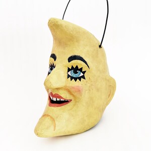 May include: A decorative, yellow crescent moon ornament with a smiling face. The moon features blue eyes with star accents, black eyebrows, and a red mouth. A black handle is attached at the top.
