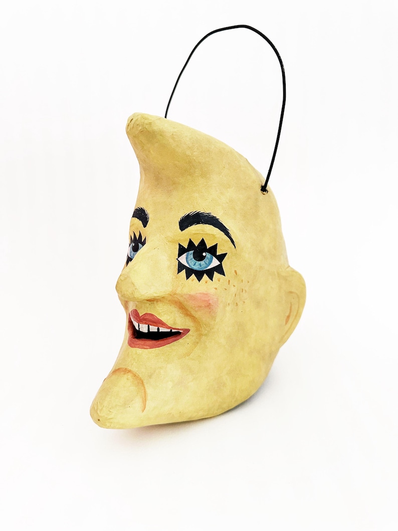 May include: A decorative, yellow crescent moon ornament with a smiling face. The moon features blue eyes with star accents, black eyebrows, and a red mouth. A black handle is attached at the top.