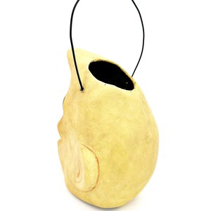 May include: A light yellow, skull-shaped container with a black handle. The container has a large opening at the top and a carved facial detail on the side. The handle is made of a thin, black wire.