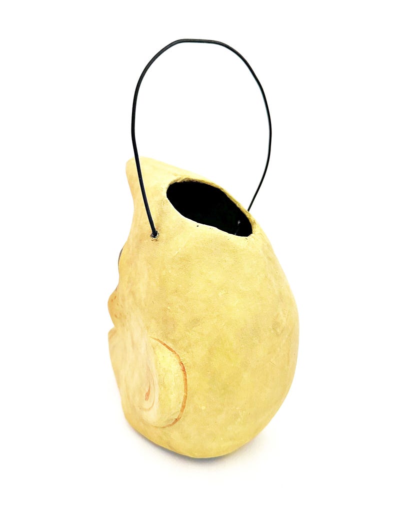 May include: A light yellow, skull-shaped container with a black handle. The container has a large opening at the top and a carved facial detail on the side. The handle is made of a thin, black wire.