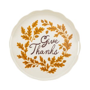 May include: A round, off-white serving tray with a scalloped edge. The center features a dark brown script text that reads "Give Thanks," surrounded by a circular wreath illustration of orange and gold oak leaves and small acorns.