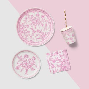 May include: A set of pink and white floral patterned party supplies, including two plates, a cup with a gold striped straw, and a napkin. The plates are round, and the cup is cylindrical. The napkin is square.