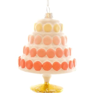 May include: A tiered cake ornament with a white base and colorful accents. The cake features layers of white frosting with orange, pink, and yellow circular decorations. It has a gold-colored base and is suspended by a silver chain.