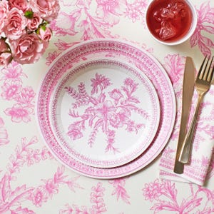 May include: Two white paper plates with a pink floral design and a pink patterned border. The plates are on a white tablecloth with a pink floral pattern. A gold fork and knife are next to a pink napkin. A glass of red liquid is in a white cup.