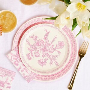 May include: A set of pink floral paper plates and napkins with a gold fork and a glass of iced tea. The plates have a cream background with a pink floral design and a pink border. The napkins have a matching floral pattern.