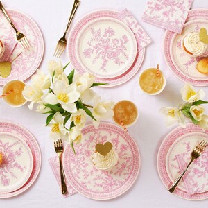 May include: A table setting with pink floral patterned plates, napkins, and gold forks. White cupcakes with gold heart toppers and glasses of orange juice with striped straws are arranged around a bouquet of white flowers.