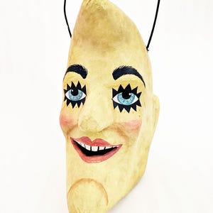 May include: A crescent moon mask with a smiling face. The mask has blue eyes, black eyelashes, and rosy cheeks. A black string is attached for hanging. The mask is painted in shades of yellow and beige.