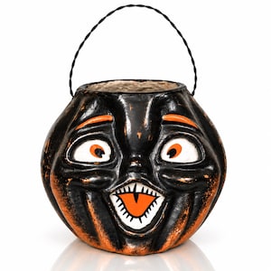 May include: A black and orange pumpkin-shaped bucket with a carved face featuring wide eyes, arched eyebrows, and a toothy grin. The bucket has a black twisted wire handle attached to the top, and the surface shows a distressed paint finish.