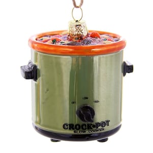 May include: A ceramic Christmas ornament shaped like a green Crock-Pot slow cooker. The ornament has an orange rim and a black control knob. The text "CROCK-POT SLOW-COOKER" is printed on the front. The ornament is suspended by a brown string.