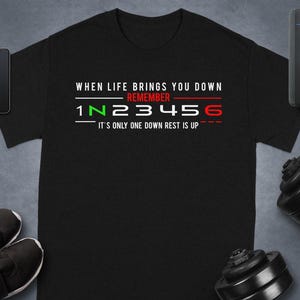 May include: Black t-shirt with the text "WHEN LIFE BRINGS YOU DOWN REMEMBER 1 N 2 3 4 5 6 It's only one down rest is up" in white, green, and red. The shirt is displayed with a smartphone, smartwatch, shoes, and weights.