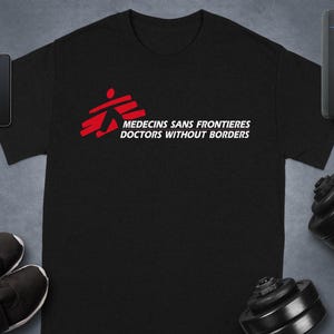 Könnte beinhalten: Schwarzes T-Shirt mit dem Logo von Ärzte ohne Grenzen in Rot und Weiß. Der Text "MEDECINS SANS FRONTIERES DOCTORS WITHOUT BORDERS" ist auf dem T-Shirt aufgedruckt. Weitere Gegenstände im Bild sind ein Smartphone, eine Smartwatch, Schuhe und eine Hantel.