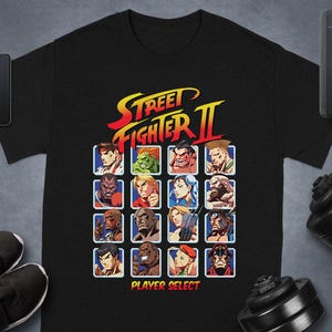 May include: Black t-shirt with a vibrant graphic of Street Fighter II characters. The design includes the game's logo and the words "Player Select". The shirt is a classic crew neck style.