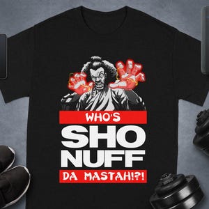 May include: Black t-shirt featuring a graphic of a character with red glowing hands and the text "WHO'S SHO NUFF DA MASTAH?!" The design is in white and red. The shirt is displayed with workout equipment and accessories.