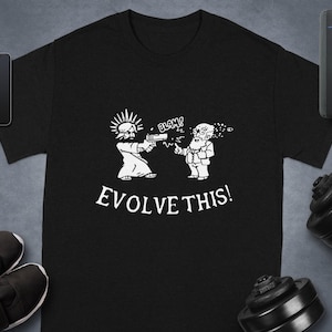Puede incluir: Camiseta negra con un diseño blanco de Jesús apuntando con una pistola a Charles Darwin. El texto "EVOLVE THIS!" está debajo. También se ven un teléfono, un reloj inteligente, un altavoz y pesas.