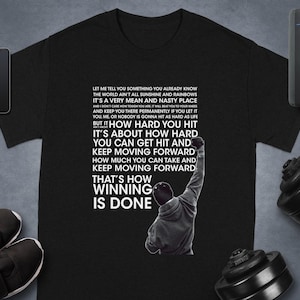 Puede incluir: Camiseta negra con una cita motivacional y la silueta de una persona levantando el puño. El texto dice: "Let me tell you something... That's how winning is done." Accesorios: reloj inteligente, teléfono y zapatillas.