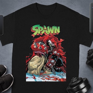 Könnte beinhalten: Schwarzes T-Shirt mit einer Comic-Illustration von Spawn. Die Figur hat einen roten Umhang, stachelige Panzerhandschuhe und ein schädelartiges Gesicht. Das Wort "SPAWN" steht in grünen und roten Buchstaben über der Figur.