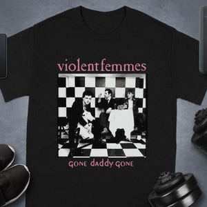 May include: Black t-shirt featuring the band Violent Femmes. The shirt has a black and white image of the band members with the text "gone daddy gone" below the image. The band's name is printed above the image in pink.