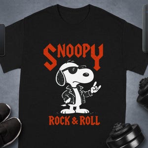 May include: Black t-shirt featuring a graphic of Snoopy in a leather jacket and sunglasses, with the text "Snoopy Rock & Roll" in red. The cartoon dog is giving a rock and roll hand gesture.