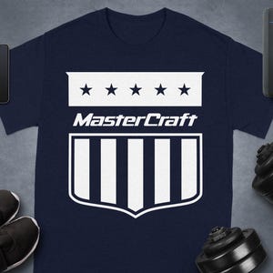 May include: Navy blue t-shirt with a white graphic design. The design features a shield shape with vertical stripes, stars, and the text "MasterCraft". Other items in the image include a smartphone, smartwatch, shoes, and weights.