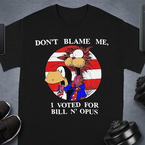 Pode incluir: Camiseta preta com o texto "DON'T BLAME ME, I VOTED FOR BILL N' OPUS." A camiseta apresenta uma ilustração de desenho animado de um pinguim e uma criatura felina dentro de um círculo vermelho, branco e azul.