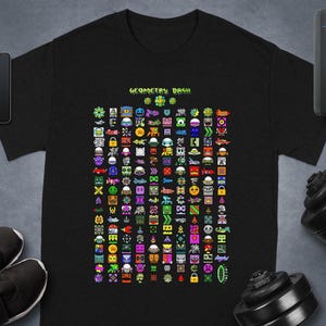 May include: Black t-shirt featuring a colorful pixelated design with various shapes and characters. The text "GEOMETRY DASH" is displayed above the design. The shirt is displayed on a gray surface.