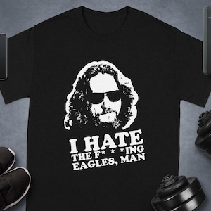 May include: Black t-shirt with a white graphic of a man wearing sunglasses and the text "I HATE THE F***ING EAGLES, MAN." The shirt is displayed with a smartphone, smartwatch, shoes, a speaker, and a dumbbell.