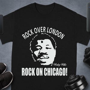 May include: Black t-shirt with a white graphic of a person's face and the text "ROCK OVER LONDON" above the face and "ROCK ON CHICAGO!" below. The text "Wesley Willis" is below the face. Accessories include a phone, watch, shoes, and a speaker.