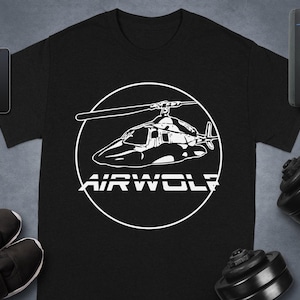 Puede incluir: Camiseta negra con un gráfico blanco de un helicóptero dentro de un círculo blanco, con la palabra "AIRWOLF" debajo. La camiseta se muestra con un smartphone, un smartwatch, zapatos y una mancuerna.