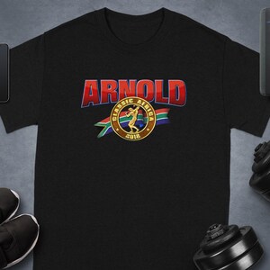 Peut inclure: T-shirt noir avec le texte "ARNOLD" en rouge et bleu, au-dessus d'un emblème circulaire avec un haltérophile et les mots "CLASSIC AFRICA 2018". Le t-shirt est entouré d'équipements et d'accessoires de fitness.