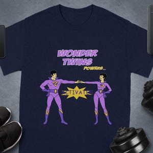 May include: Navy blue t-shirt featuring a cartoon illustration of two figures in purple superhero costumes, with the text "WONDER TWINS POWERS... ACTIVATE!!" in purple and yellow. The design is centered on the shirt.