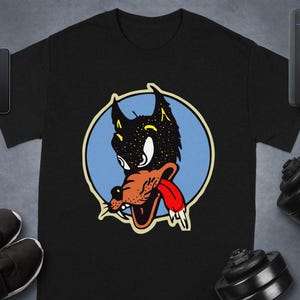 May include: Black t-shirt featuring a cartoon wolf head with a starry sky design inside the head, a red tongue, and a blue circle background. The wolf has a brown snout and is outlined in yellow. The shirt is displayed on a gray surface.