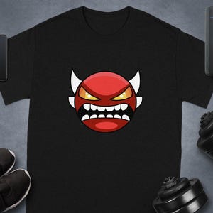 Puede incluir: Camiseta negra con una cara de diablo de dibujos animados roja, cuernos blancos, ojos amarillos y una boca muy abierta con dientes blancos. La camiseta está sobre una superficie gris con un smartphone, smartwatch, zapatos y pesas.