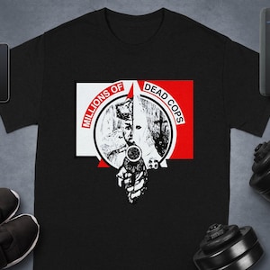 May include: Black t-shirt with a graphic design. The design includes the text "MILLIONS OF DEAD COPS" in red and white, and an illustration of a person holding a gun. The image is surrounded by a smartphone, a smartwatch, a speaker, a dumbbell, and trainers.
