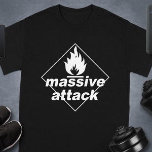 Puede incluir: Camiseta negra con un gráfico blanco de una llama dentro de un rombo y las palabras "massive attack" debajo. La camiseta se muestra con un smartphone, un reloj inteligente, zapatos y una pesa.