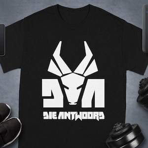 Könnte beinhalten: Schwarzes T-Shirt mit einem weißen Grafikdesign eines stilisierten Tierkopfes und dem Text "DIE ANTWOORD". Das T-Shirt wird auf einer dunklen Oberfläche mit Sportgeräten und Zubehör präsentiert.
