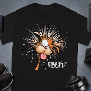 Puede incluir: Camiseta negra con un diseño de cara de gato de dibujos animados. El gato tiene pelaje naranja, ojos blancos y una lengua rosa, con el texto "THBBFT!" en blanco. La camiseta se muestra con equipos de fitness y accesorios.