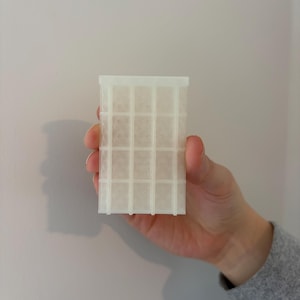 May include: A translucent, rectangular object with a grid pattern is held in a hand. The object appears to be made of a light-colored, possibly plastic material. The grid is composed of vertical and horizontal lines, creating a series of small squares.