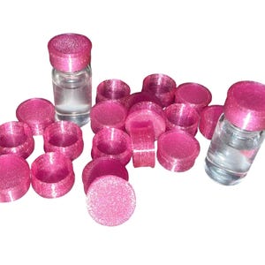 May include: A collection of small, round, bright pink glittery containers with matching lids, scattered alongside two clear glass vials topped with identical pink glittery caps. The containers are empty, while the two glass vials contain liquid.
