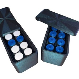 May include: Two rectangular dark teal storage cases with hinged lids featuring a starburst pattern. The left case contains eight small vials with white caps, while the right case holds eight small vials with blue caps, arranged in two rows.